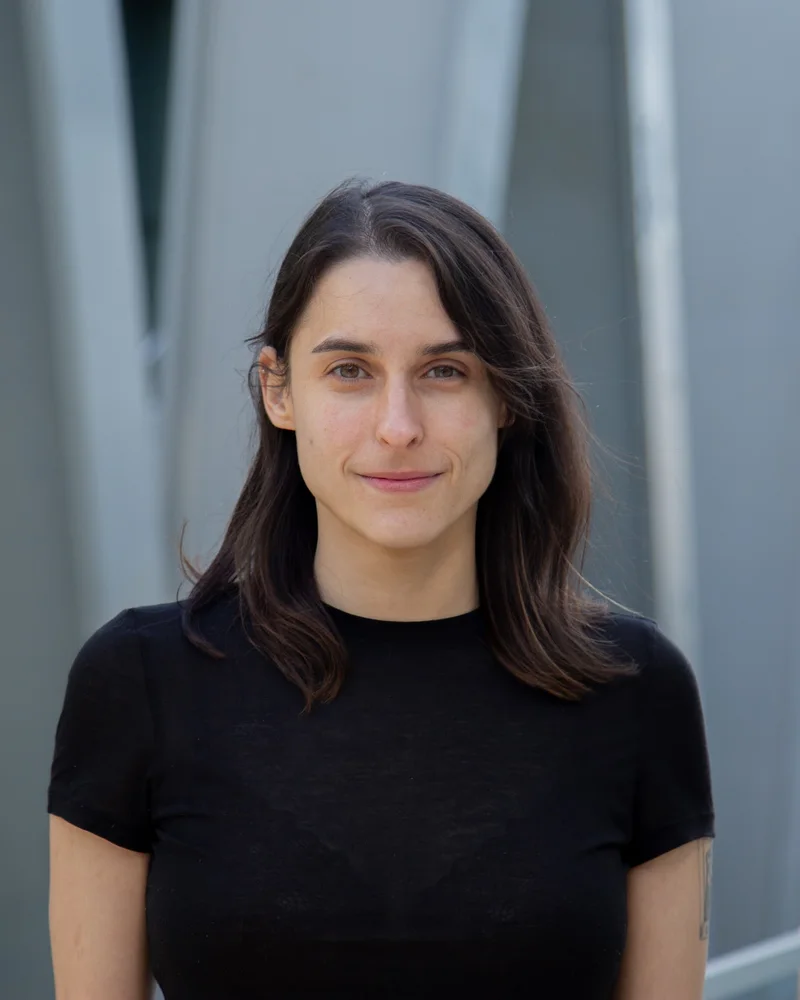 Grazia Prato, smiling, in a black T-shirt in front of a grey building.